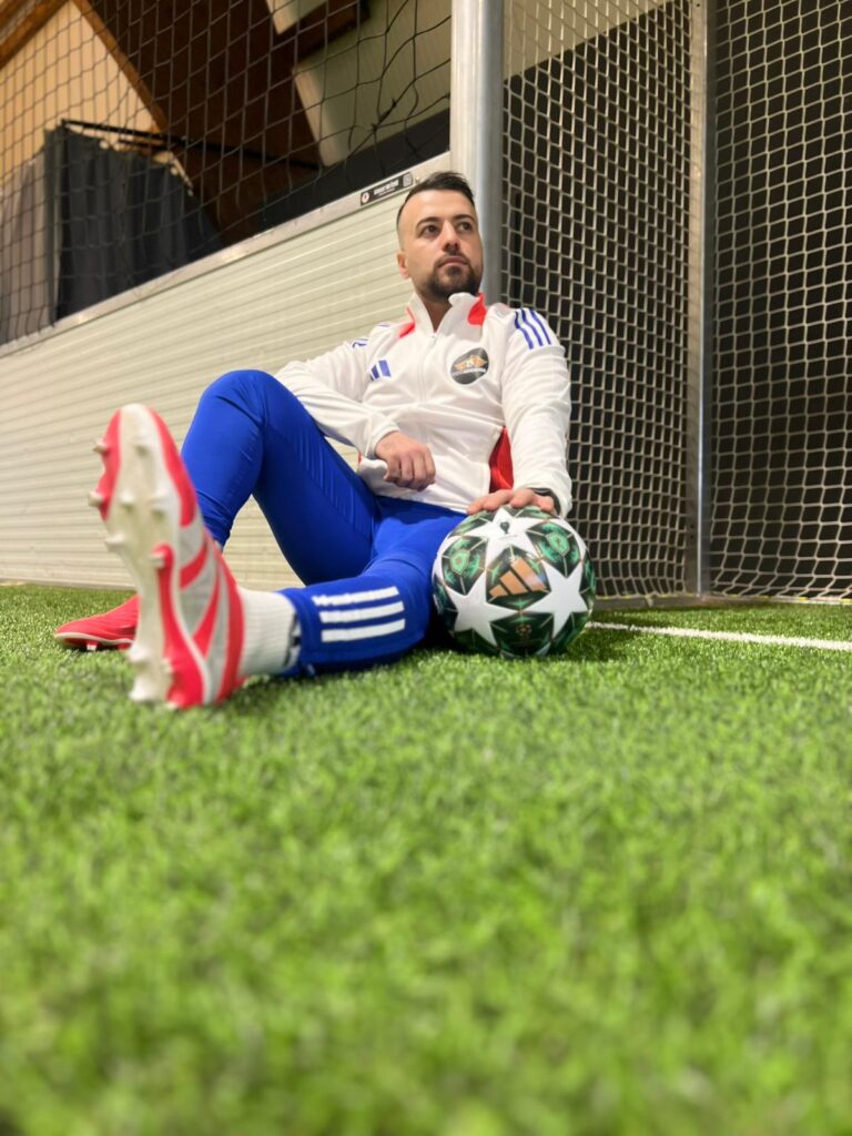 Ein Mann mit dunklem Bart sitzt nachdenklich im weißen Trainingsanzug am Fußballtor auf grünem Kunstrasen und hält einen bunten Fußball