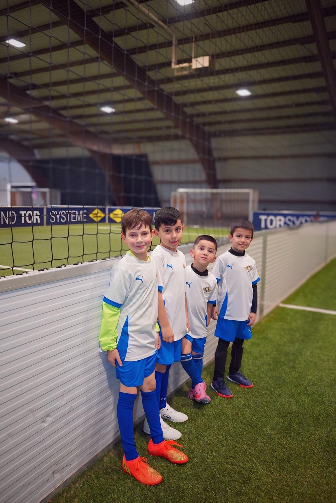 Vier junge Jungen im weißen Fußballtrikot lächeln nebeneinander am Hallenrand
