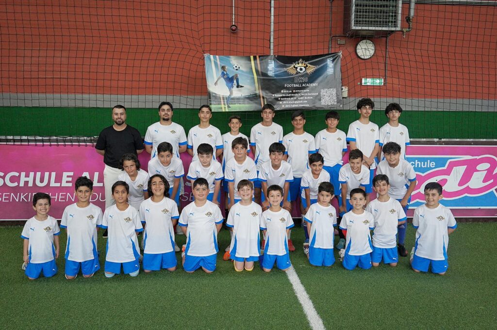 Eine Gruppe lachender Jungen im weißen Trikot mit blauem Rand posiert mit ihrem Trainer auf einem Kunstrasenfeld