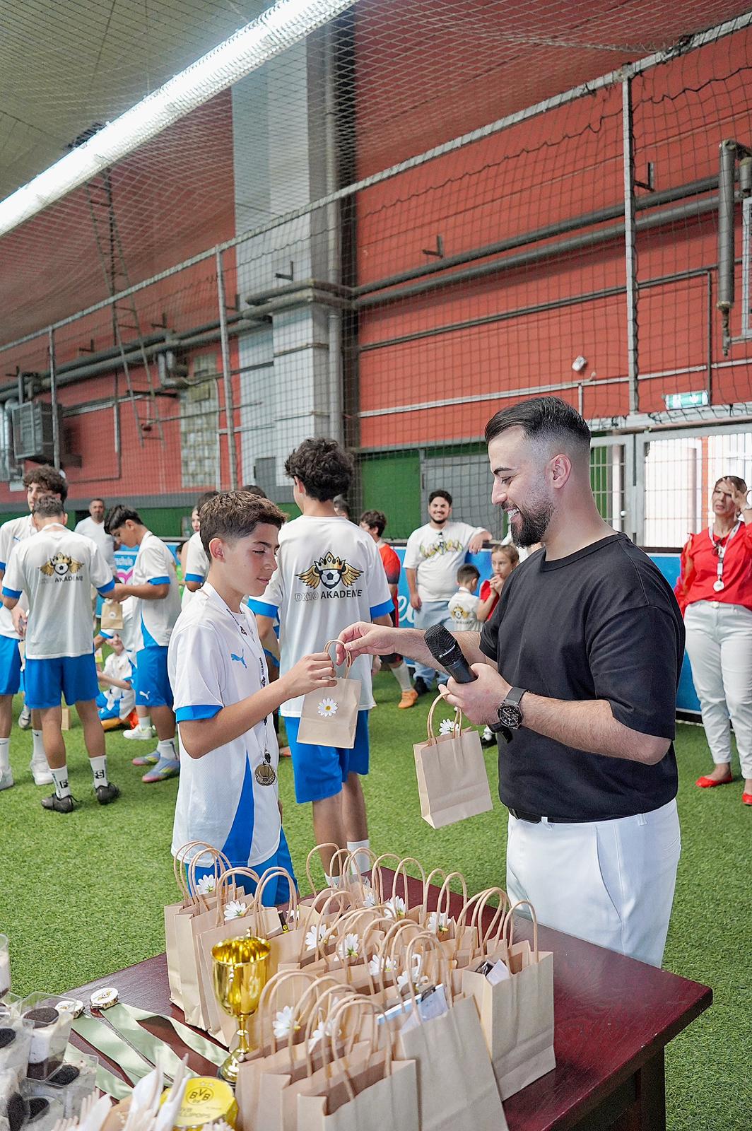 Ein Mann mit Bart und schwarzem Shirt überreicht einem Jungen ein Geschenk auf grünem Kunstrasen
