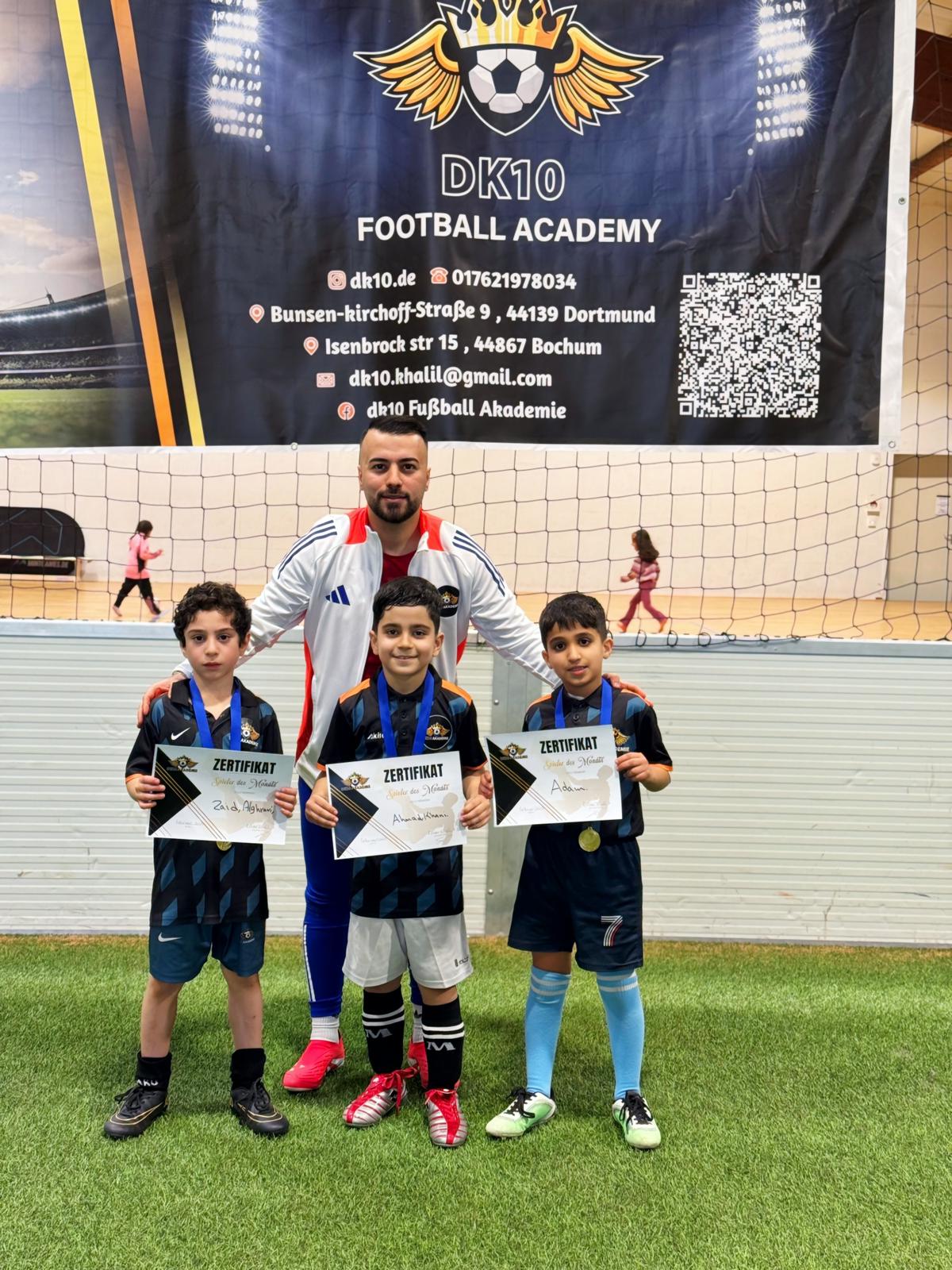 Ein Trainer mit Bart steht lachend hinter drei Jungs im Fußballtrikot, die stolz Zertifikate halten