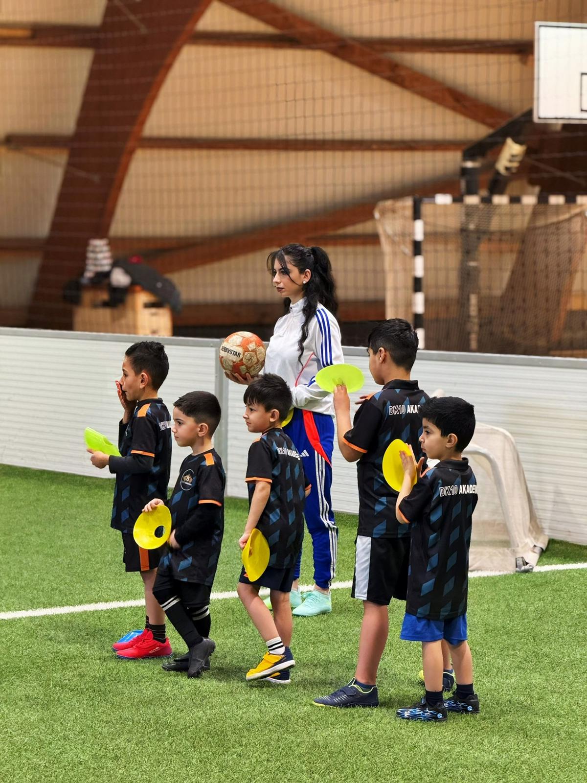 Eine junge Frau mit Ball lächelt freundlich und beaufsichtigt sechs spielende Kinder