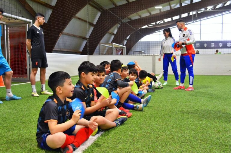 Kinder sitzen lächelnd in einer Reihe in bunter Sportkleidung auf grünem Kunstrasen